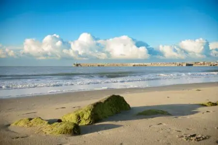 A beach with a large stone in front of the sea with a harbour arm in the background