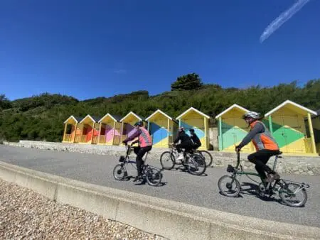 Four people with helmets cycle past colourfully painted beach huts on a promenade