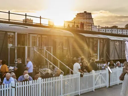 People enjoy drinks by a white picket fence and in front of fixed railway carriages