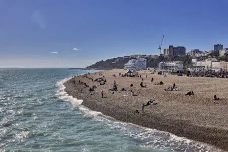 People sit or stand on a shingle beach with large buildings in the background