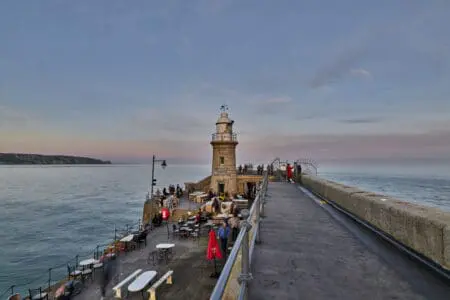 A lighthouse stands at the end of a harbour arm with people on the chair and tables in front of it