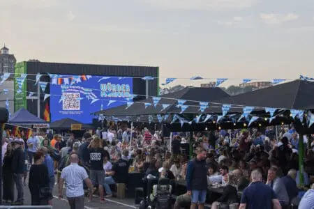 A large crowd of people socialise in front of a big screen covered by black awnings