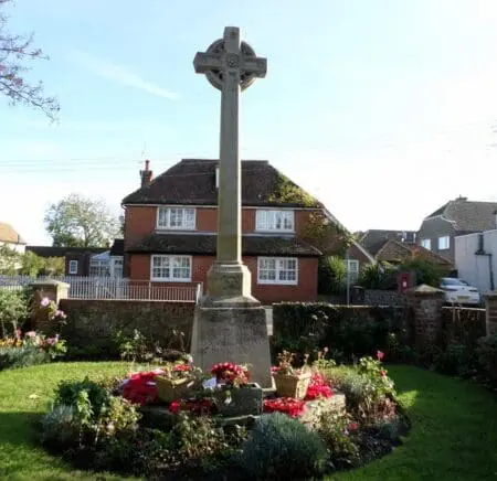 A stone cross on a plinth is surrounded by a flowerbed and then grass in a residential street.
