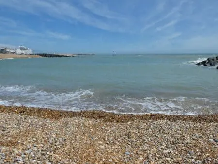 A shingle bay under a blue sky with white buildings in the background.
