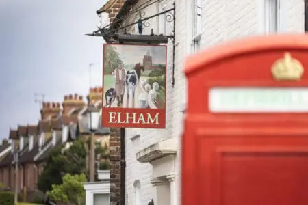 A pub style sign which says Elham with a picture of a man herding sheep and a cow. There is a red postbox in the foreground of the picture and a row of houses behind