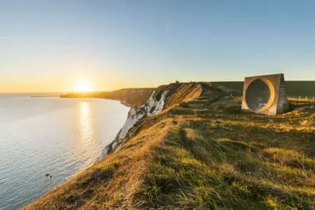 A sound mirror stands on green grass above white cliffs and out to a blue sea