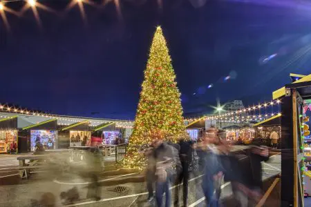 Blurred people moving in front of a large Christmas tree and huts selling goods