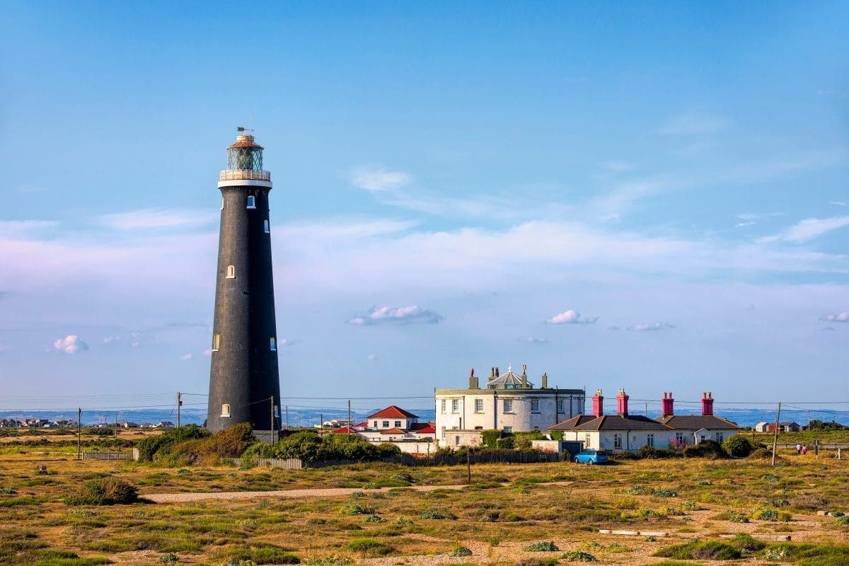 A black lighthouse sits on the horizon next to a few houses in the foreground is grass scrubland.