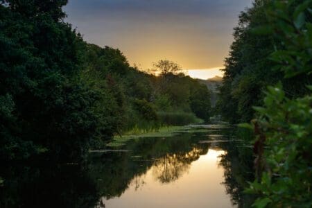 A canal stretches into the distance bordered by mature trees and with a sunset in the distance