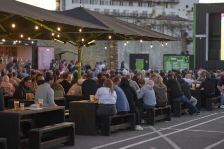 A large crowd of people sit together on benches under a black awning