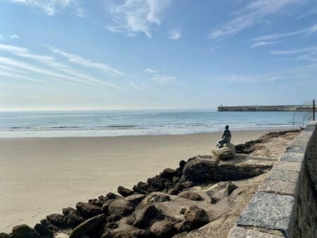 A rocky wall with a statue of a mermaid beside a sandy beach on a sunny day