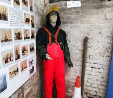A mannequin in orange waders stands near a traffic cone and beside a board of pictures and words