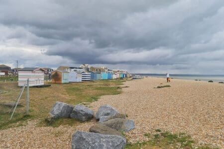 A shingle beach lined by colourful beach huts on the left and the sea on the right with a man walking across it.