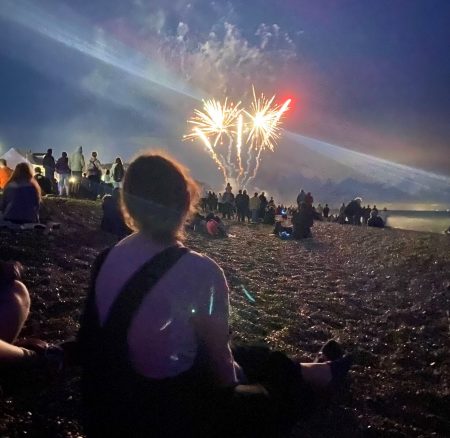 A woman sits on a shingle beach in the evening watching a fireworks with a crowd